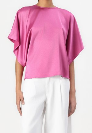Femme portant un chemisier rose brillant, ample, à manches courtes larges, et un pantalon blanc plissé taille haute.