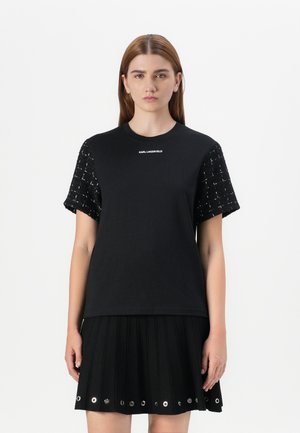 A young woman models a black top with textured sleeves, paired with a pleated black skirt featuring decorative grommets.