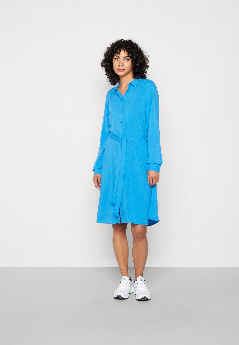 Vestito camicia blu con maniche lunghe, chiusura a bottoni e cintura da annodare. Tessuto morbido, lunghezza fino al ginocchio, con orlo leggermente curvo.