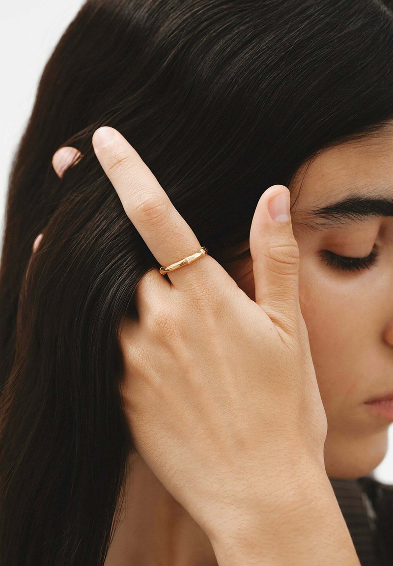 Mujer con los ojos cerrados y largo cabello oscuro metiéndose el cabello detrás de la oreja, llevando un anillo de oro simple en su dedo medio.