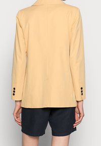 Blazer giallo chiaro con una vestibilità su misura, spacco centrale sul retro e dettagli con bottoni neri sulle maniche, abbinato a pantaloni corti scuri.