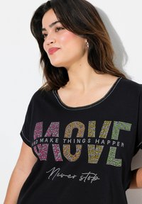 Camiseta negra de manga corta, con texto colorido de rhinestones que dice "MUEVE Y HAZ QUE LAS COSAS SUCEDAN" y un cuello de contraste.
