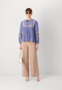 Blåmønstret bluse med en løs pasform og samlet detaljering, parret med beige vidde bukser og strop sandaler. Creme håndtaske.