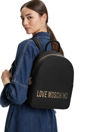 Una giovane donna con una giacca di jeans blu porta sulla spalla uno zaino nero Love Moschino con scritte dorate e cerniera.