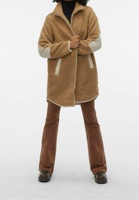 Cappotto in pile marrone con colletto, dettagli crema sulle maniche e sulle tasche, zip frontale e vestibilità comoda, abbinato a pantaloni marroni svasati e stivaletti scuri.