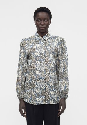 Personne à la peau foncée portant une chemise boutonnée à manches longues avec un motif floral bleu, blanc et beige, debout devant un fond blanc uni.