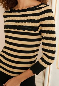 Maglione a righe nere e beige con scollatura a onda e lavorazione a maglia testurizzata. Le maniche lunghe presentano polsini a coste, accentuando il design.