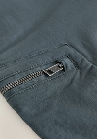 Tamno siva tkanina s teksturiranom površinom; sadrži vidljivi metalni zip i bočnu džep; dizajnirana za korisnost i praktičnost.