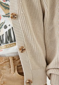 Beige gebreide cardigan met een gestructureerd patroon, voorzien van bruine knopen. Gedragen over een graphic t-shirt in aardetinten met een casual pasvorm.