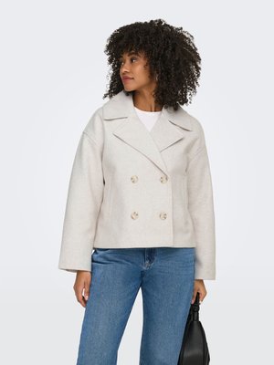 Sieviete ar cirtainiem matiem, valkājot gaiši bēšu dubultpogas mēteli, zilas džinsas un tur melnu rokassomiņu, skatās uz kreiso pusi.