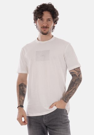 Hombre con cabello rizado y tatuajes en ambos brazos, vistiendo una camiseta blanca y vaqueros grises, posando con una mano en el bolsillo.