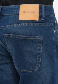 Jeans de denim azul con una etiqueta de cuero con "ONLY & SONS" grabada. Bolsillo trasero visible y detalles de costura. Corte clásico.