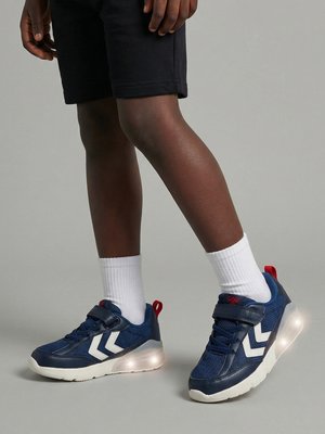 Barn iført marineblå sneakers med hvide chevroner og lysende såler, hvide sokker og sorte shorts mod en grå baggrund.