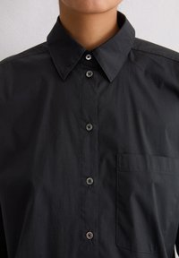 Persoon die een zwart knoopshirt draagt met een puntige kraag en een borstzak, zichtbaar van nek tot halverwege de tors.