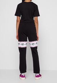 Svart t-shirt och joggers har en vit och rosa remsa med ögmönster vid låren. Joggersarna har en avslappnad passform. Vita sneakers med lila detaljer.