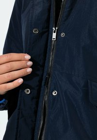 Tummansininen, kevyt takki, jossa on sileä, kiiltävä pinta; edessä vetoketju ja nepparikiinnitykset; käsi lepää vetoketjun lähellä.