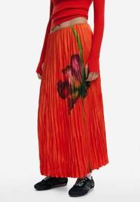 Jupe plissée rouge ornée d'un grand motif floral coloré. Fabriquée dans un matériau brillant, elle possède une silhouette fluide et une taille élastique.
