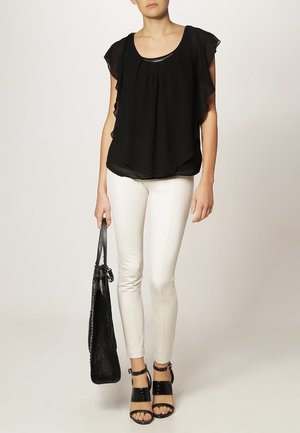 Vrouw met een zwarte blouse met wappermouwen, witte strakke broek, zwarte sandalen met hakken en een zwarte handtas in haar hand.