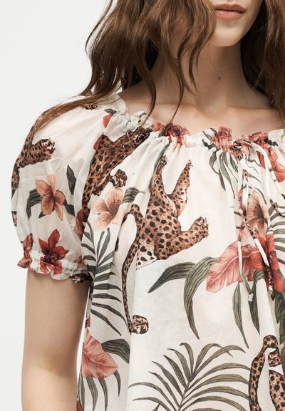 Γυναίκα με φούστα off-shoulder με τύπωμα λεοπάρ και τροπικά λουλούδια σε αποχρώσεις του καφέ, πράσινου και κόκκινου σε λευκό ύφασμα.