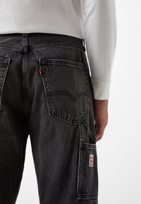 Pantalones de mezclilla gris oscuro con bolsillos traseros y una etiqueta de logo roja. Las características incluyen costuras visibles y un diseño de corte relajado. Se muestra una camiseta de manga larga blanca.