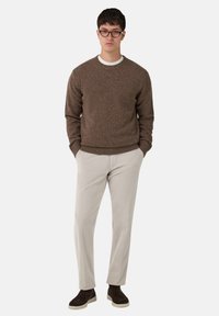 Brūns teksturēts adījuma džemperis virs baltas kreklu ar apaļu kakla izgriezumu, kombinēts ar gaiši beidzamām pieguļošām biksēm un tumši brūnām zamšādām kurpēm.