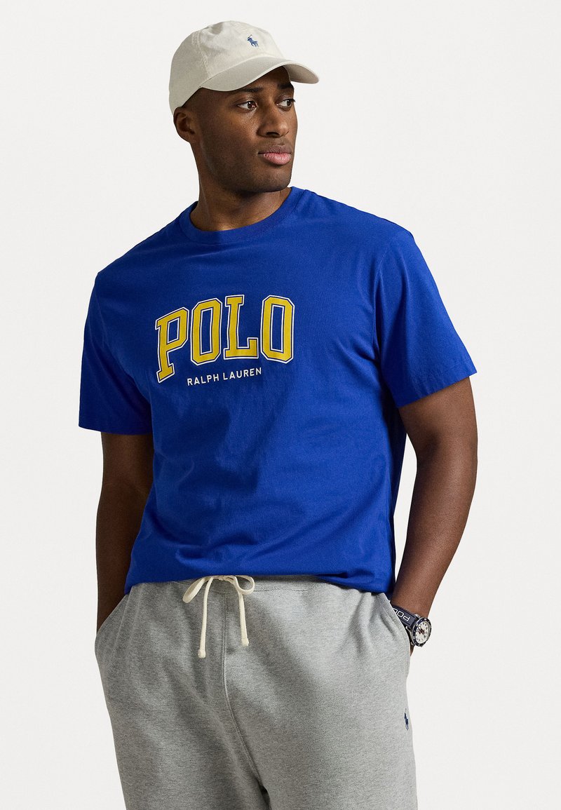 Royal blue t-shirt with "POLO RALPH LAUREN" in yellow text. Paired with light gray sweatpants and a cream cap. Notable watch detail visible.