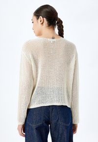 Cremfarbener, locker gestrickter Pullover mit langen Ärmeln und abgerundetem Saum. Leichte Textur mit lockerer Passform, kombiniert mit dunkelblauen Jeans.