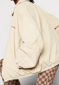 Béžová kordová bunda s límcem, objemovými rukávy, elastickými manžetami a vyšívaným textem na jedné straně. Spárováno s kostičkovanými kalhotami.