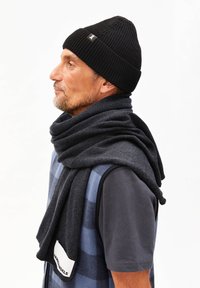 Un bonnet en tricot noir et une écharpe gris foncé drapés sur un gilet à carreaux bleus et un t-shirt gris ; la vue de profil met en valeur la texture et les superpositions.