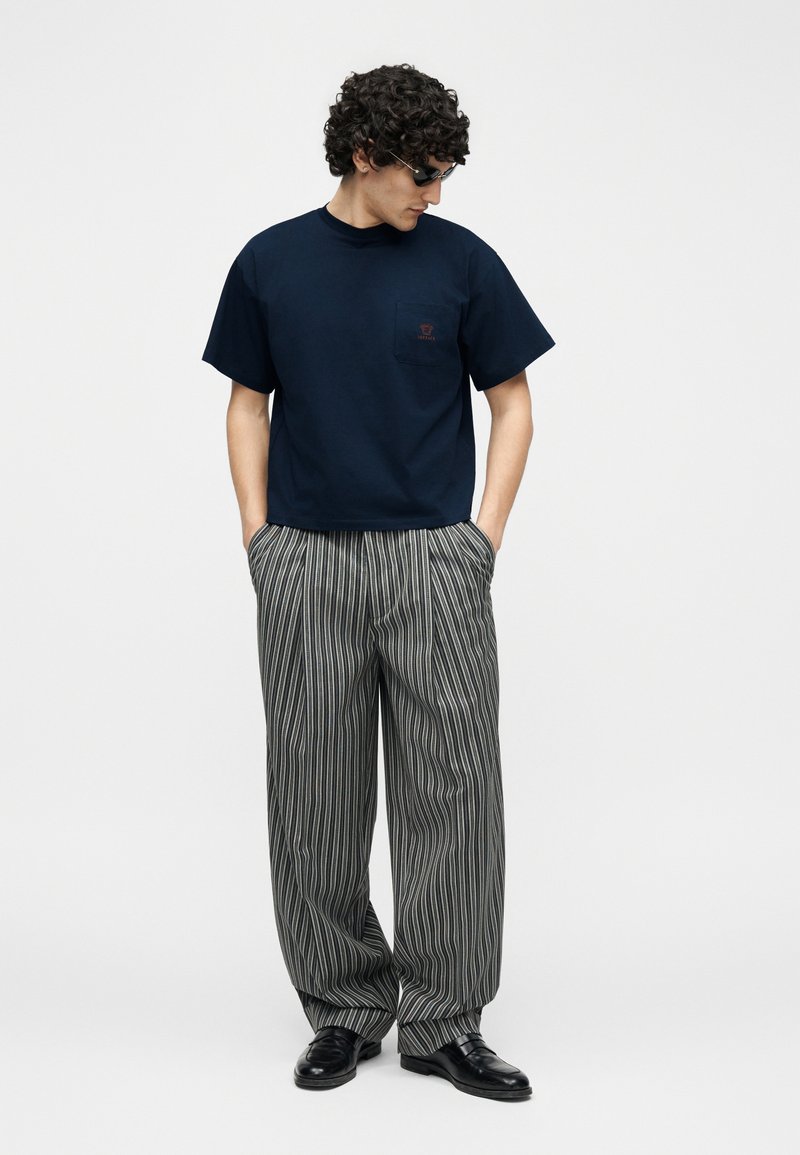 Uomo con occhiali da sole neri, maglietta a maniche corte blu navy con taschino sul petto, pantaloni larghi grigi a righe sottili e scarpe eleganti nere.