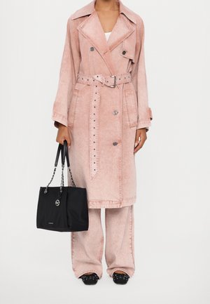 Femme portant un trench-coat long rose ceinturé et un pantalon large assorti, tenant un sac à main noir à bandoulières en chaîne, et chaussée de chaussures noires cloutées.