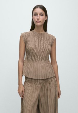 Kobieta ubrana w bezrękawnikowy beżowy plisowany top i dopasowane szerokie plisowane spodnie, stojąca na tle jednolitego jasnego tła.