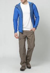 Veste légère bleue pour homme avec des accents gris, associée à un pantalon cargo beige. Les deux pièces présentent des éléments de design fonctionnels pour une utilisation en extérieur.