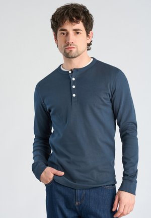 Giovane uomo con capelli castani ricci che indossa una maglietta Henley blu navy a maniche lunghe e jeans blu, in piedi con una mano in tasca.