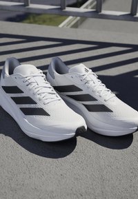 White athletic shoes with black stripes on the sides, made of textured mesh and synthetic materials, featuring a rounded toe and lace-up design.