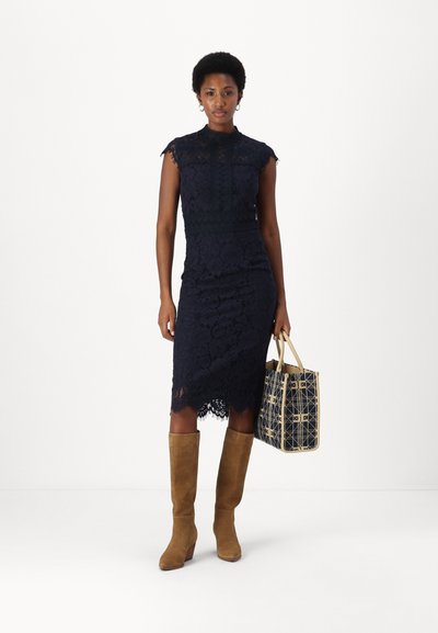 Rochie din dantelă de culoare bleumarin, până la genunchi, cu guler înalt și mâneci scurte. Botine înalte din suede maro. Ținând o geantă de umăr cu un model.