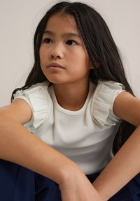 Maglietta bianca a coste con maniche arricciate, collo rotondo e trama uniforme, indossata da un bambino seduto con le braccia incrociate.