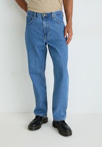 Mees, kes kannab siniseid sirgeid teksaseid ja musti nahast nööriga saapaid, seisab ühevärvilise valge tausta ees.