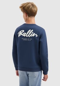 Navyblauwe sweatshirt met ribgebreide manchetten en onderrand, met witte geborduurde tekst "Ballin" en "Echoes of the Urban Wild" op de achterkant.
