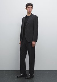 Schwarzer maßgeschneiderter Anzug mit einem strukturierten Blazer und geraden Hosen, kombiniert mit einem schwarzen, eng anliegenden Pullover und schwarzen Slippern auf einer strukturierten Oberfläche.