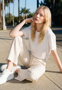 Young woman with long blonde hair sits on a sidewalk in casual white clothing and sneakers, resting her chin on her hand under clear skies.