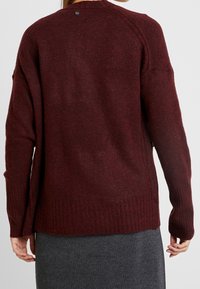 Bordowy sweter z dzianiny o teksturowym wykończeniu, z prążkowanymi mankietami i dołem oraz małym guzikiem z tyłu. Luźny krój z bocznymi szczelinami.