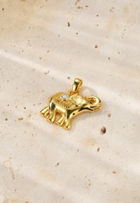 Colgante de elefante de oro con un intrincado diseño, que presenta una pequeña piedra preciosa clara, sobre un fondo de piedra texturizada clara.