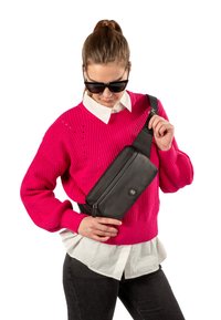 Borsa a tracolla nera con finitura lucida e chiusura con zip. Indossata sopra un maglione lavorato a maglia rosa acceso e una camicia bianca a strati, con jeans scuri.