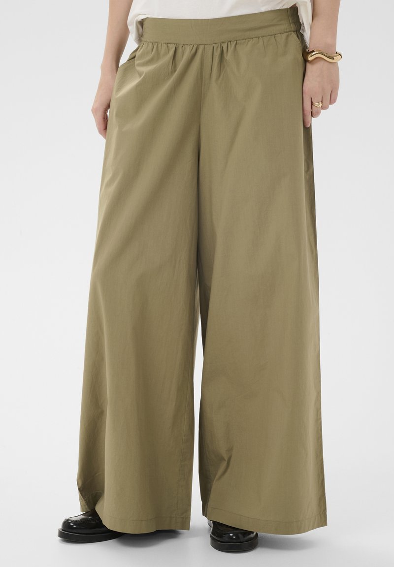 Femme portant un pantalon large vert olive avec une taille élastique et des chaussures noires brillantes, debout devant un fond uni.