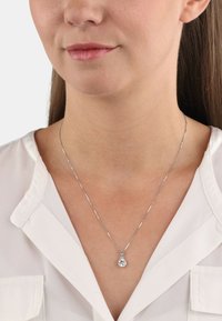 Collar de plata con un colgante en forma de gota que presenta una gema transparente. La cadena tiene un acabado texturizado, complementando una camisa blanca.