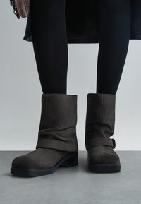 Brune sued stövletter med en vikbar kant, ett band med detalj i guldspänne och en låg svart gummihäl. Bärs med svarta leggings.