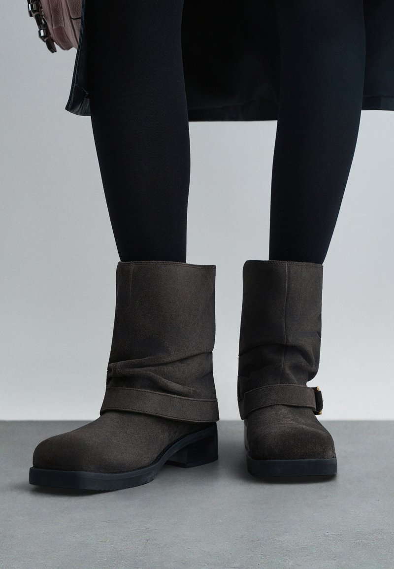 Brune sued stövletter med en vikbar kant, ett band med detalj i guldspänne och en låg svart gummihäl. Bärs med svarta leggings.