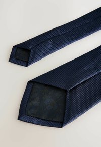 Marineblaue, strukturierte Krawatte mit einem Rautenmuster, die an der Spitze mit einem floralen Muster gefüttert ist. Das Material wirkt gewoben und glatt.