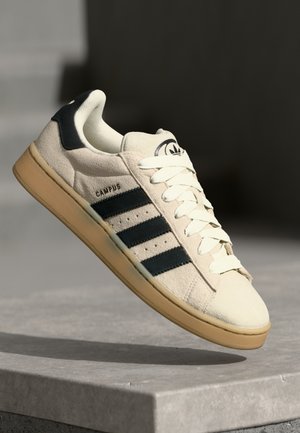Beige suede sneaker with three dark blue stripes, white laces, and a tan rubber sole. Features a logo on the tongue and "CAMPUS" text.